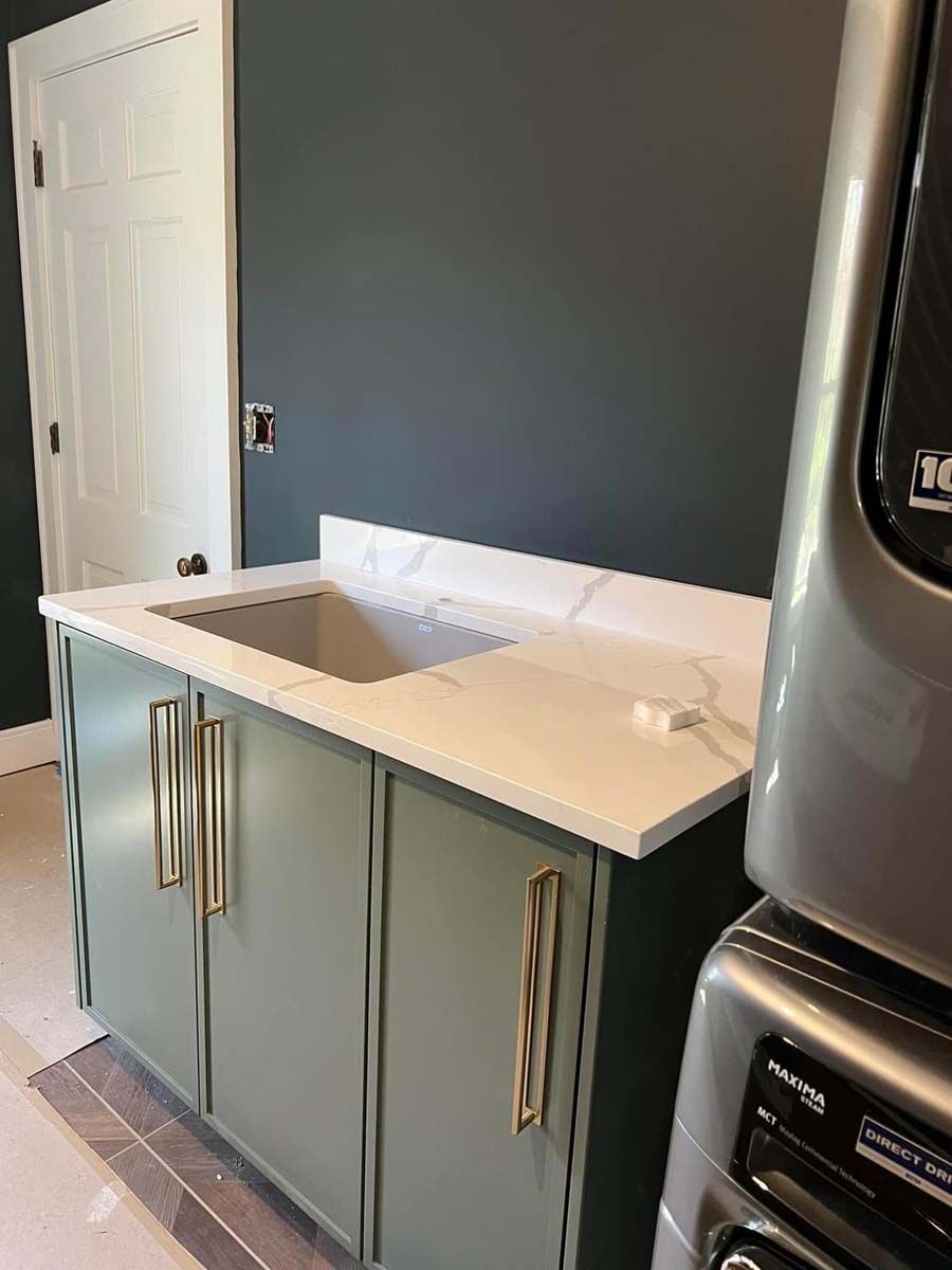 Laundry room with sage green cabinets, white countertop, and gold handles; a sink and a stacked dryer.