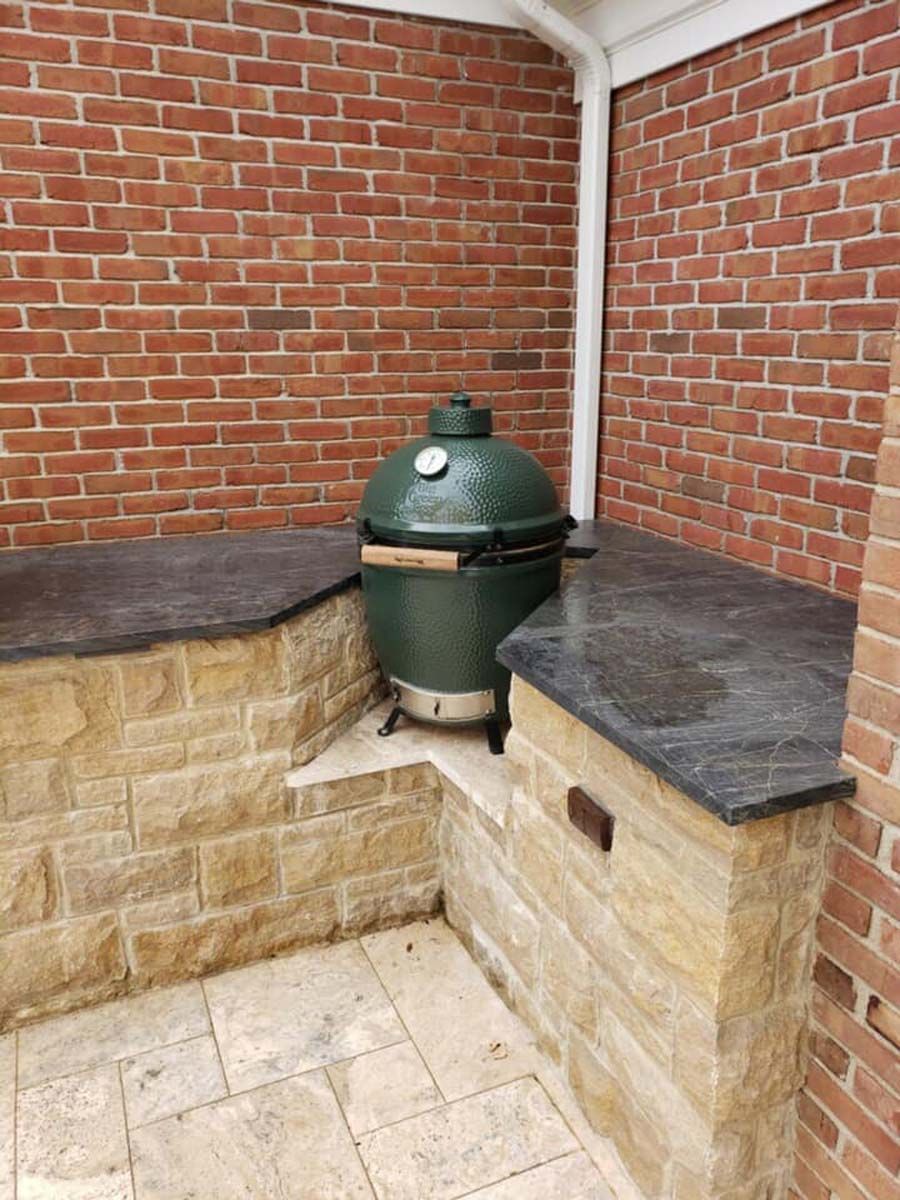 A green Big Green Egg grill on a stone counter, set in a brick and stone outdoor kitchen.