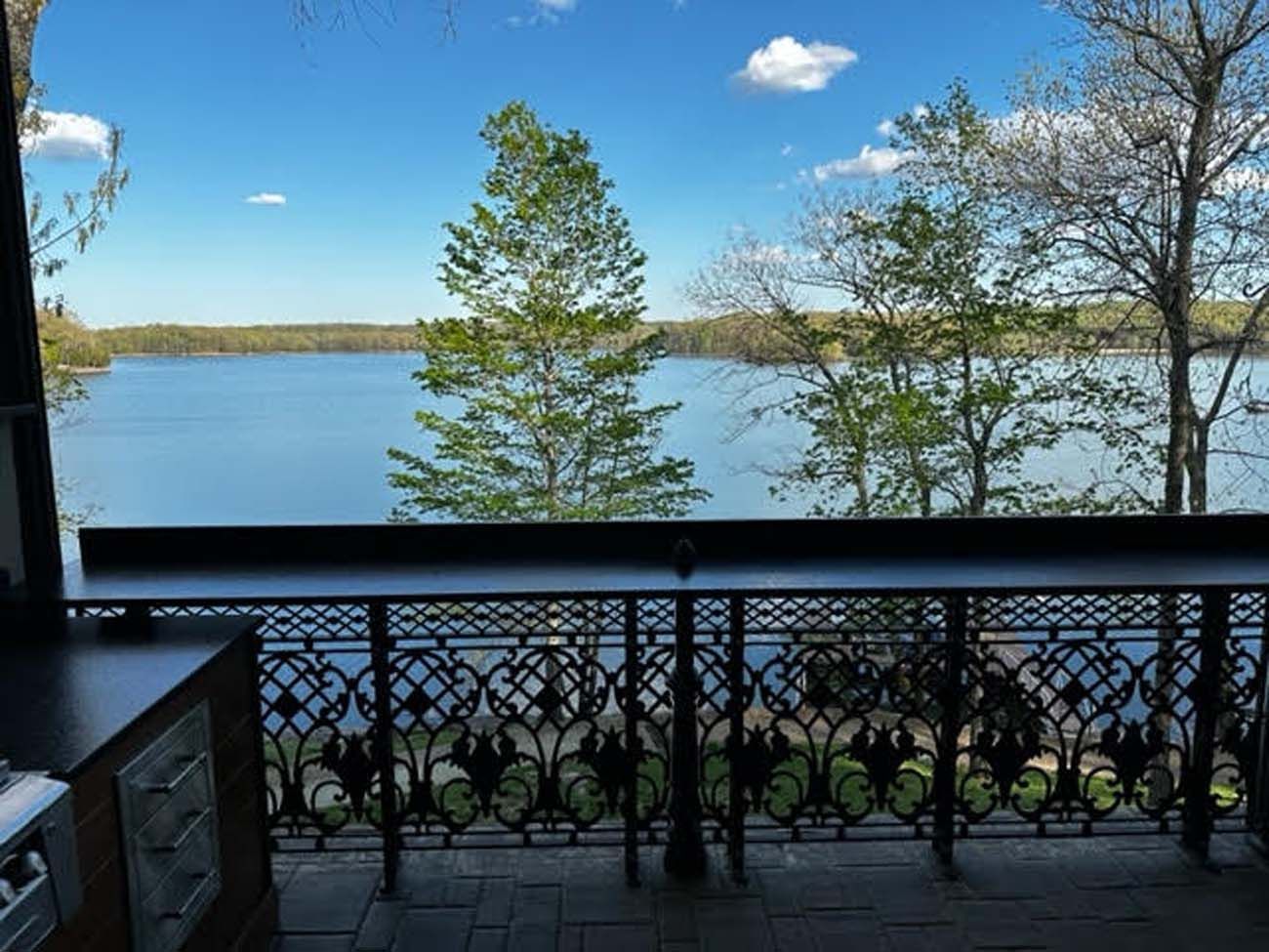 Lake view from a patio with ornate black railing, trees, and a partly cloudy blue sky.