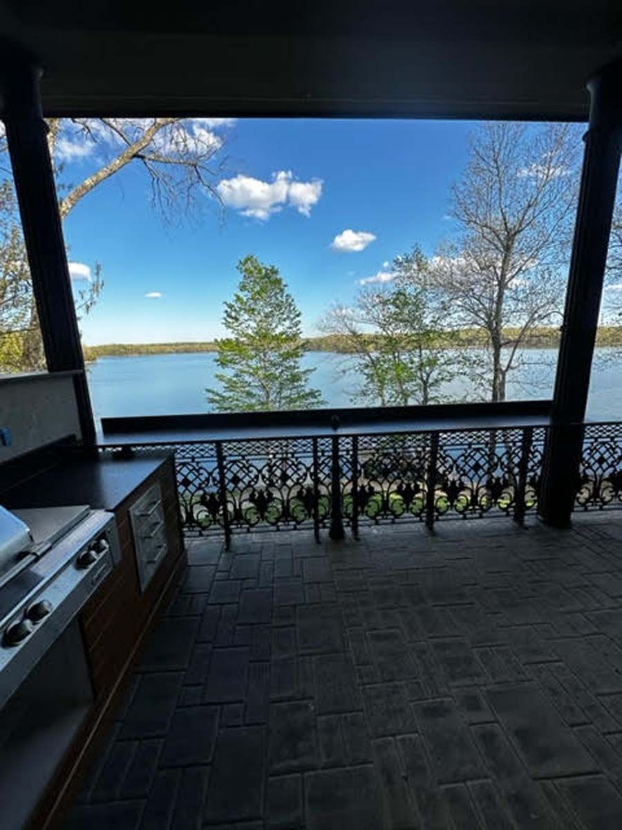 Patio overlooking a lake, with a built-in grill and decorative iron railing, under a bright blue sky.