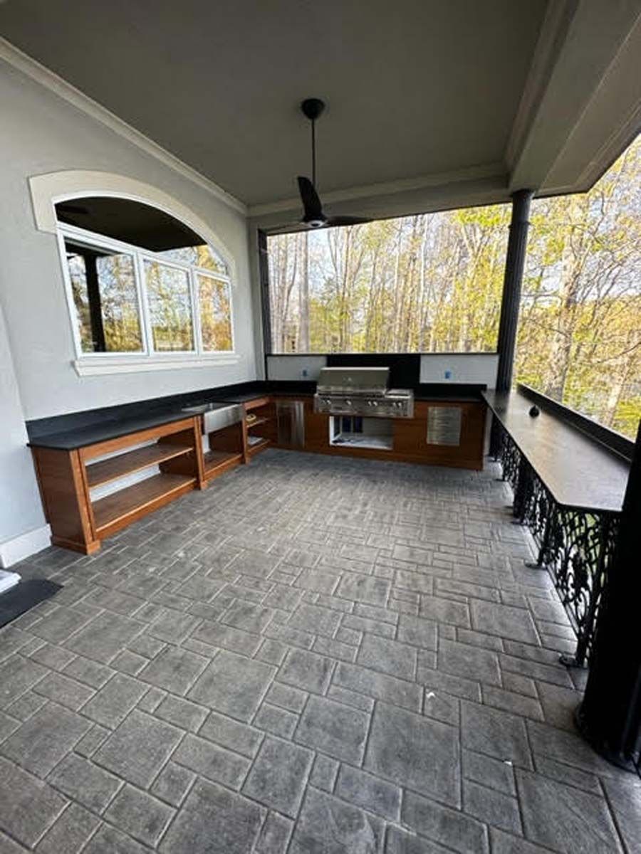 Outdoor kitchen on a covered patio with gray brick-patterned flooring, wooden cabinets, and a grill.
