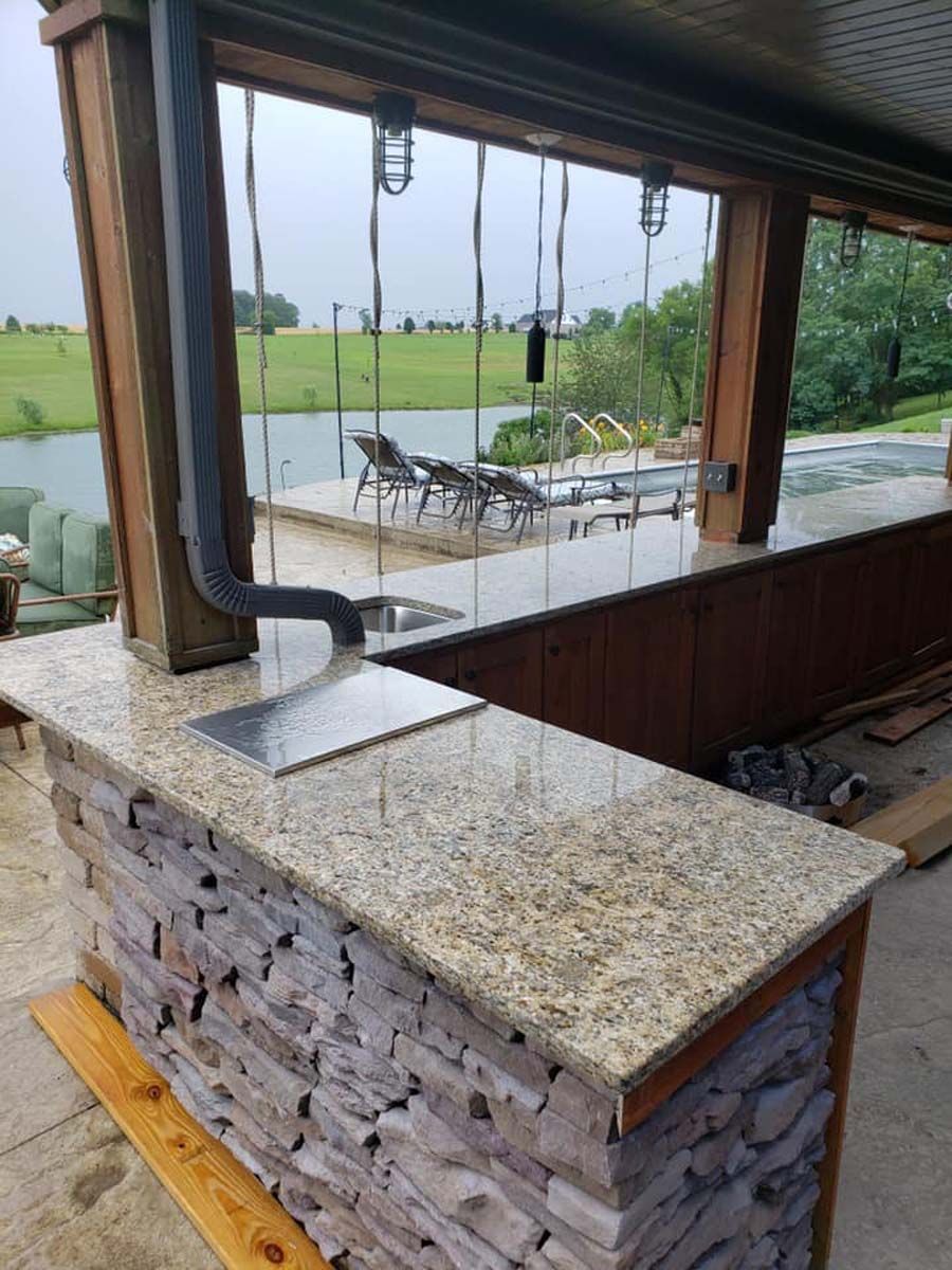 Outdoor kitchen with stone facade bar, granite countertop, wooden beams, and view of lake.