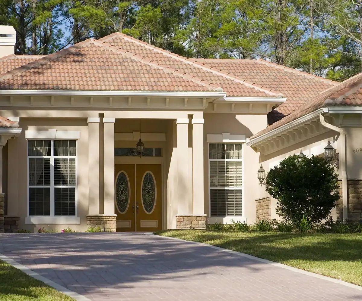 Beige stucco house with a tile roof, pillars at the entrance, and a brick driveway.