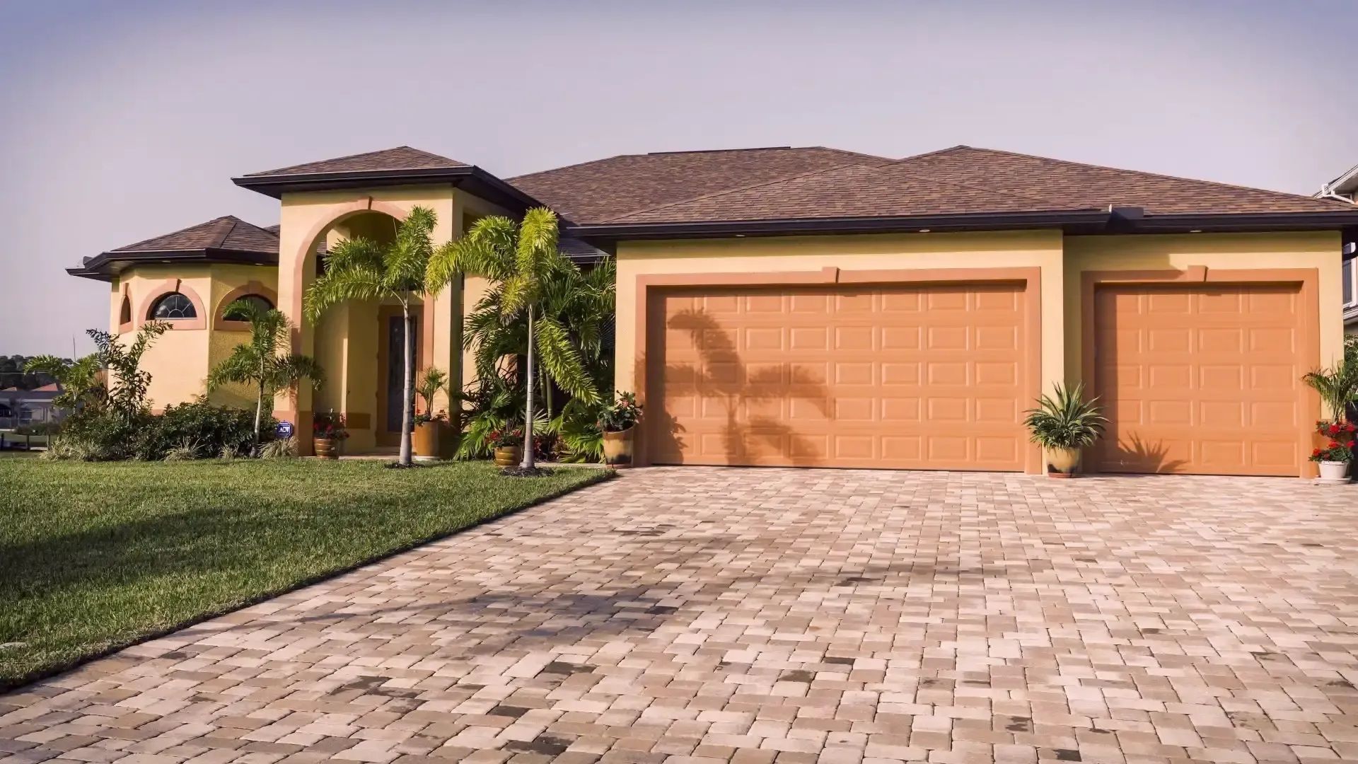 Tan house with brown roof and garage doors, brick driveway, green lawn, palm trees.