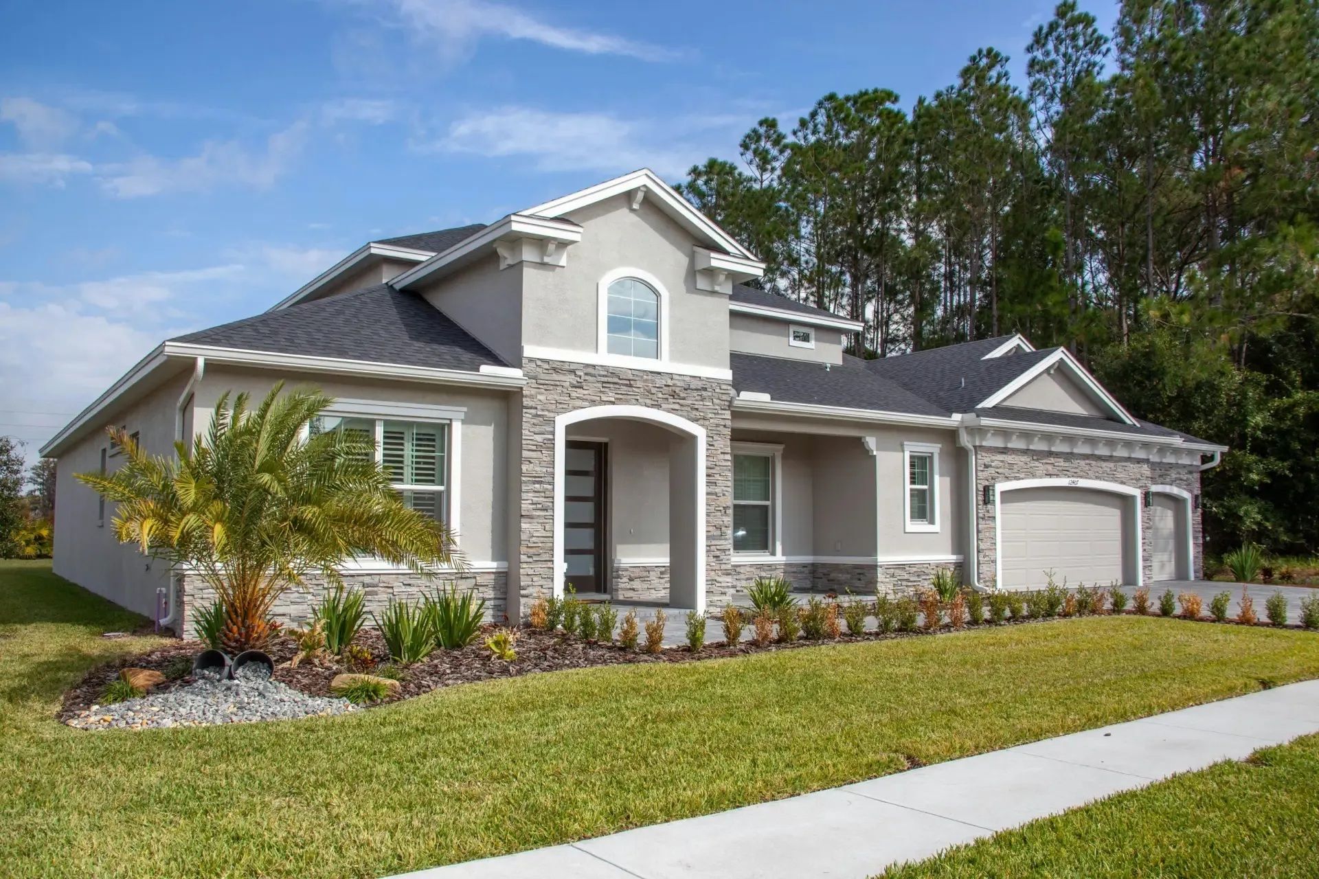 Gray stucco house with stone accents, green lawn, palm tree, and three-car garage under a blue sky.