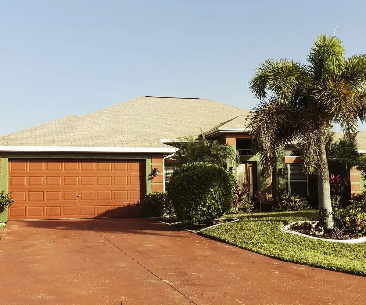 House with red garage door and driveway, palm trees and green landscaping.