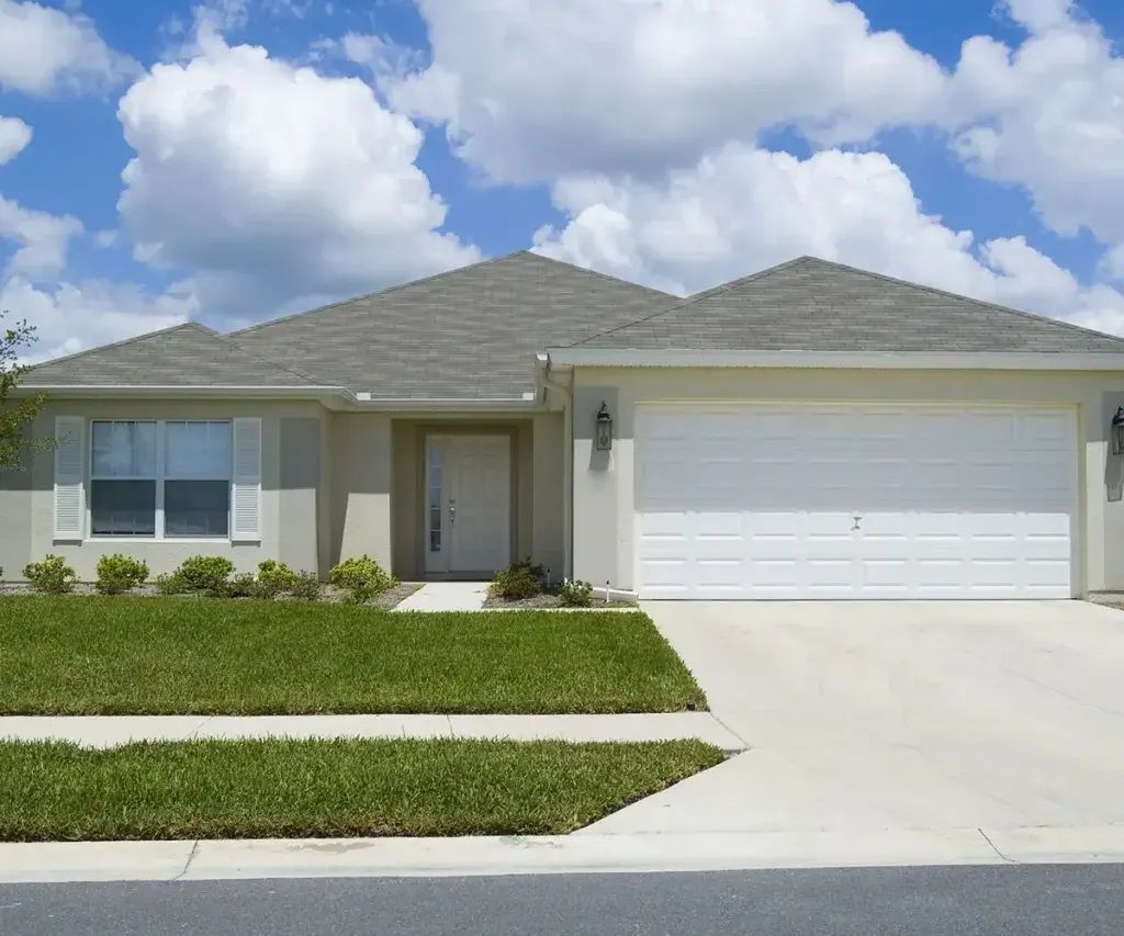A one-story beige house with a gray roof, white garage door, and green lawn under a blue sky.