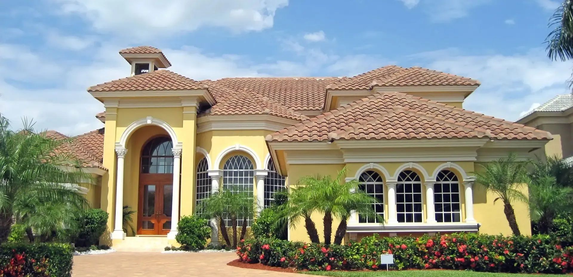 Yellow stucco house with red tile roof, arched windows, and lush landscaping on a sunny day.