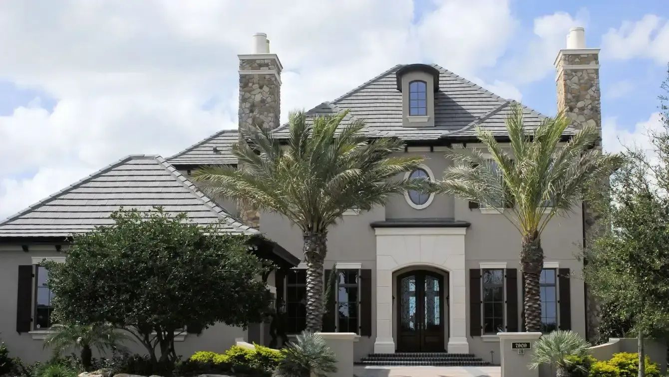 Elegant gray stucco house with dark shutters, palm trees, and stone chimneys.