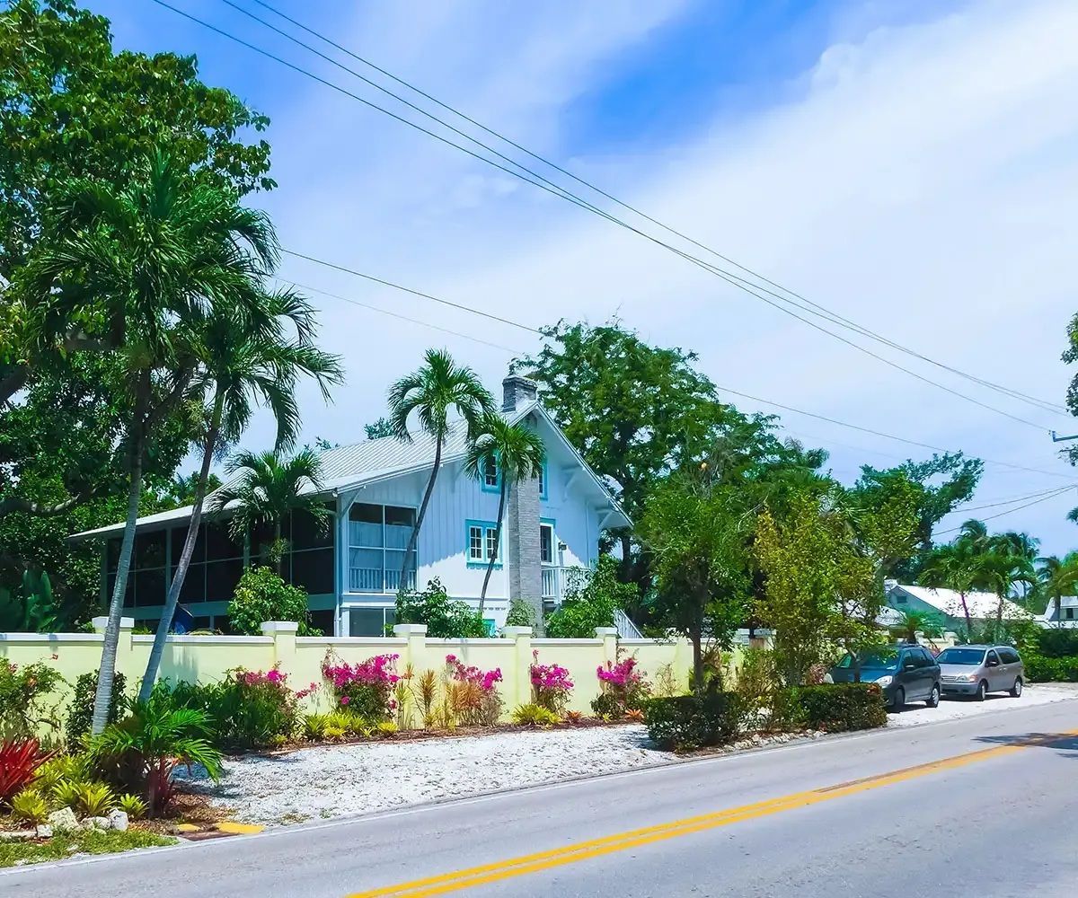 White house with blue trim behind a low wall with pink flowers, palm trees, and parked cars on a sunny street.