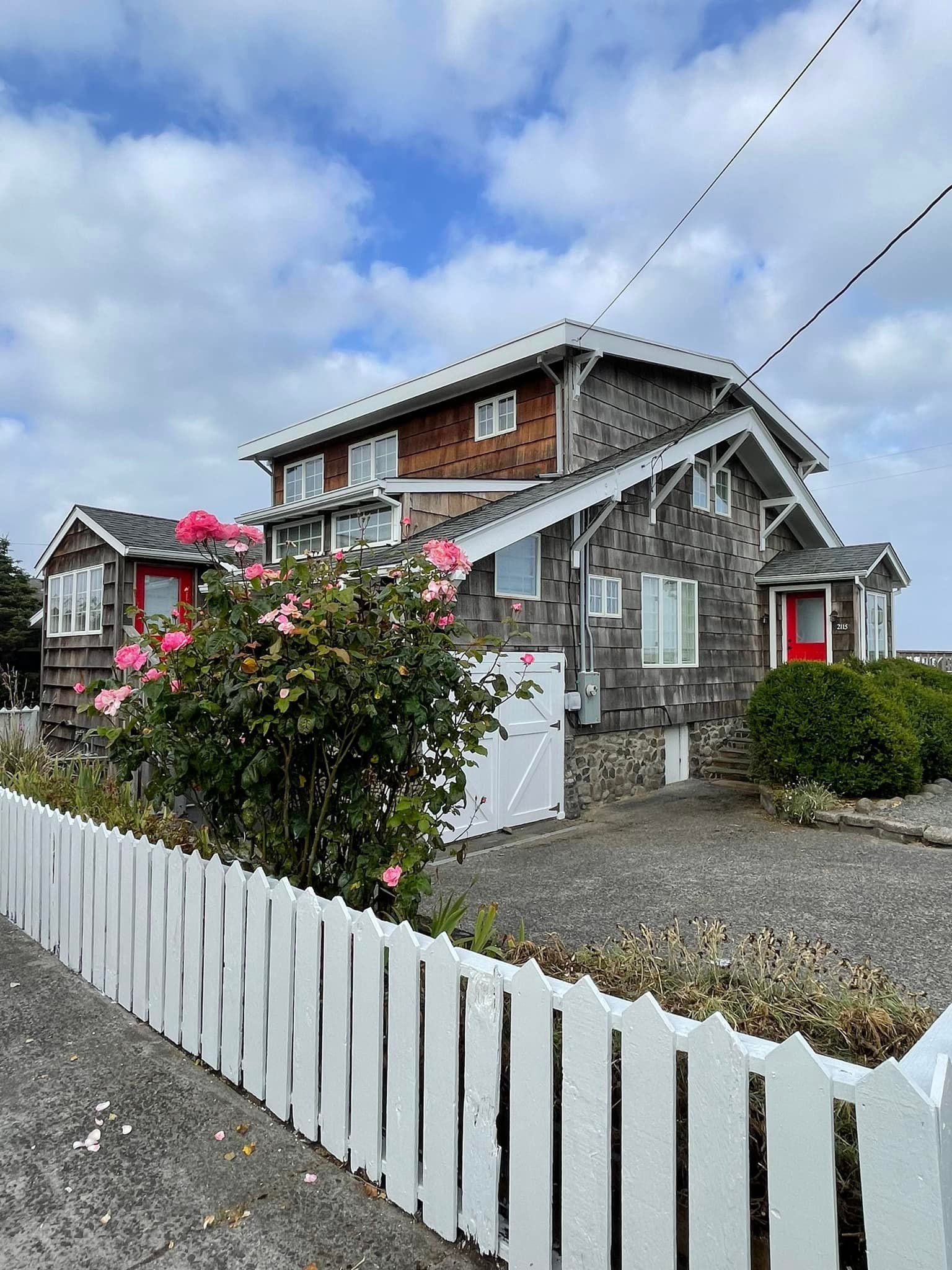 Large, weathered shingled house with red door, white picket fence, pink flowers under cloudy sky.