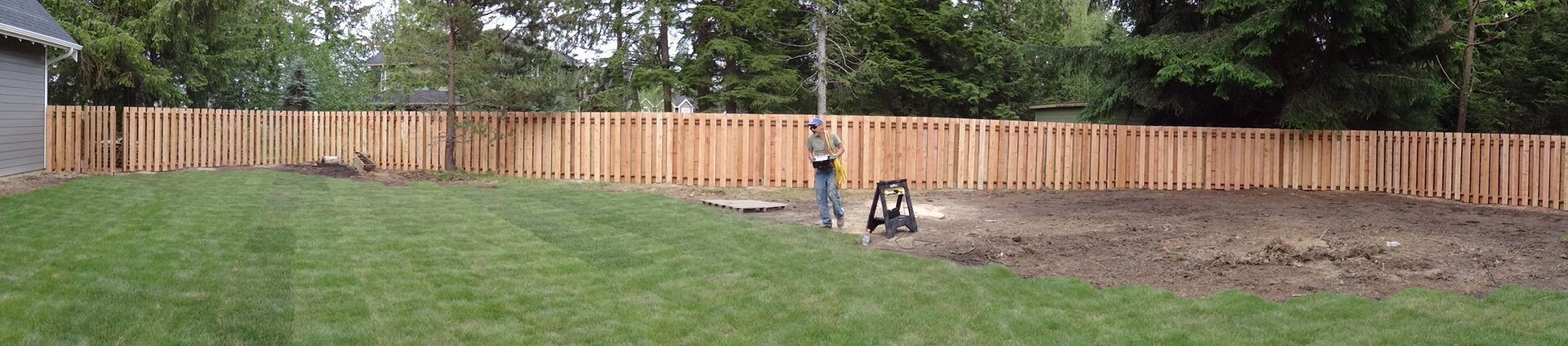 Backyard with wooden fence and a person working near a tool. Green grass and trees in the background.