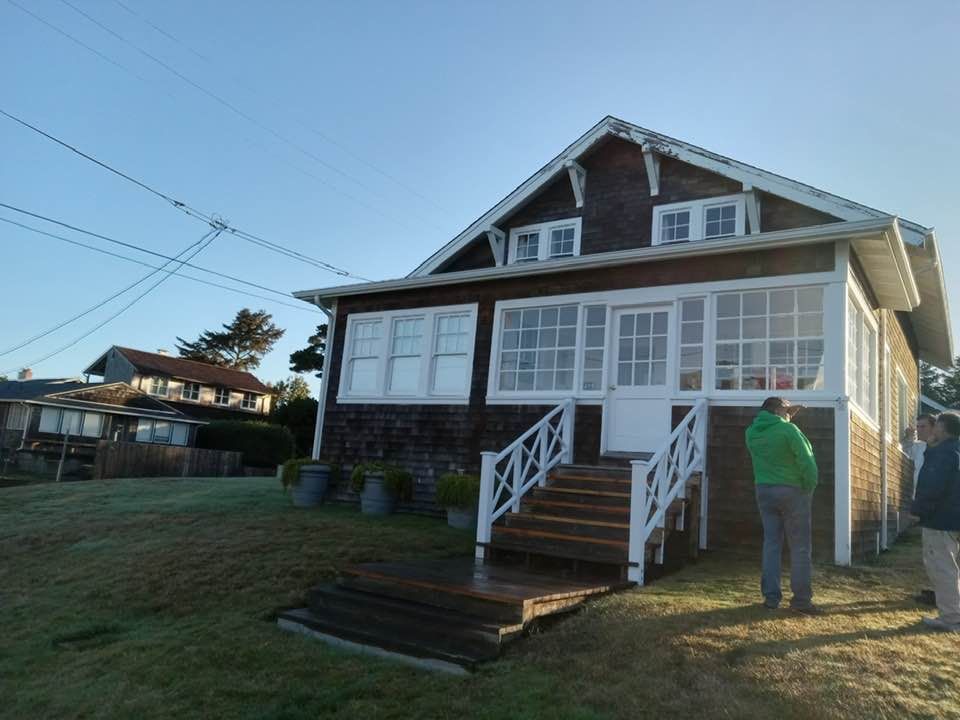 Brown shingled house with white trim and porch. Two people stand outside on a sunny day.