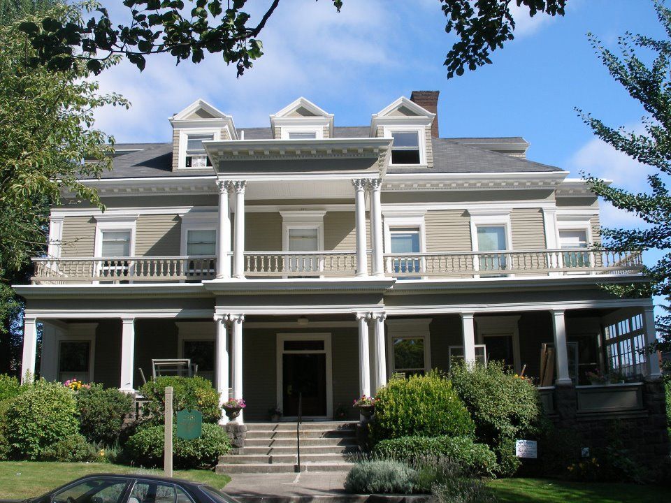 Large, two-story pale green house with white columns, balconies, and dormers; set in a yard with greenery.