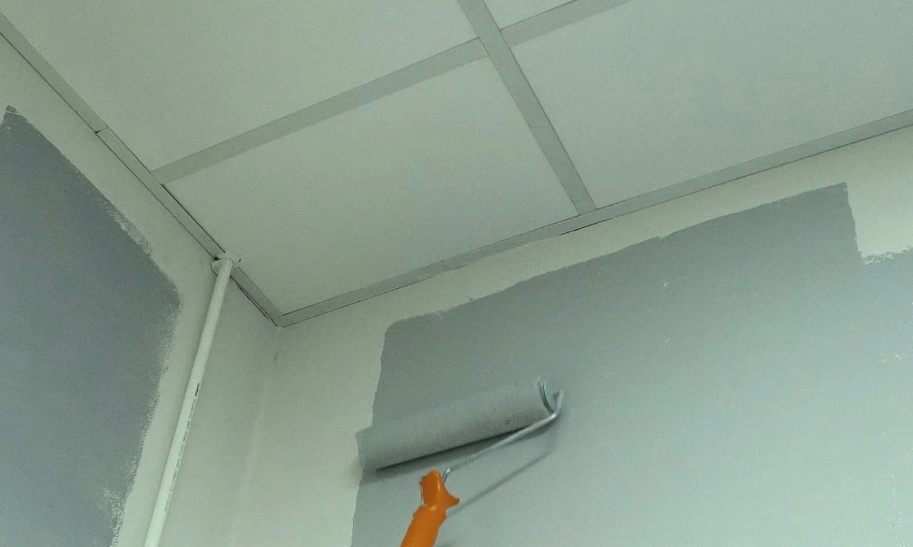 Painting a light gray wall with a roller in a corner of a room.