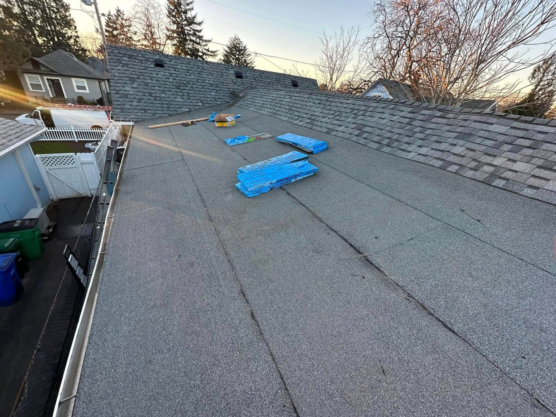 Rooftop with shingles. Blue tarp and supplies are visible. Houses and trees in the background during sunset.