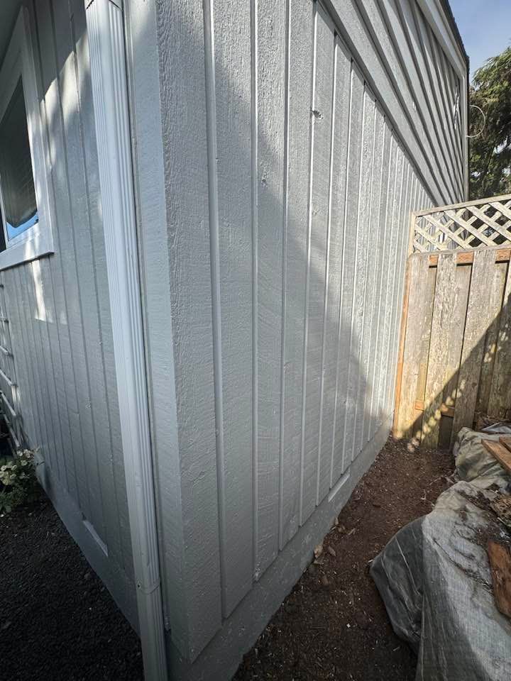 Gray siding of a building's exterior, with white trim, next to a wooden fence and a garden.