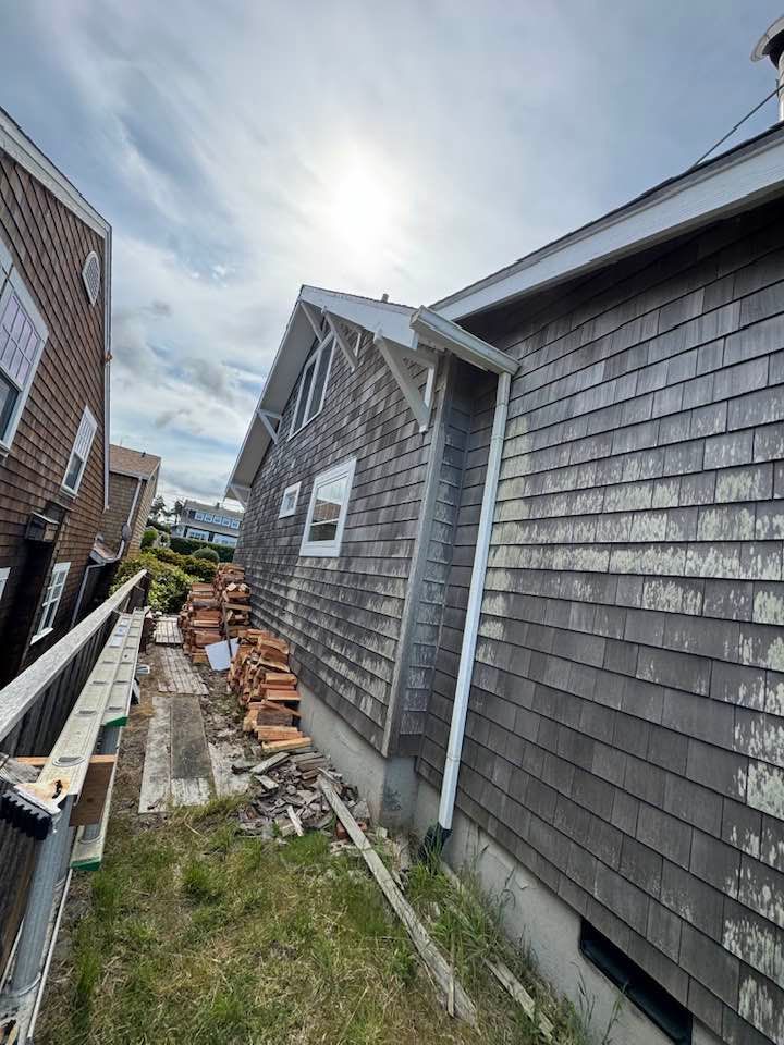 Weathered shingled house under a cloudy sky. Construction materials in yard.