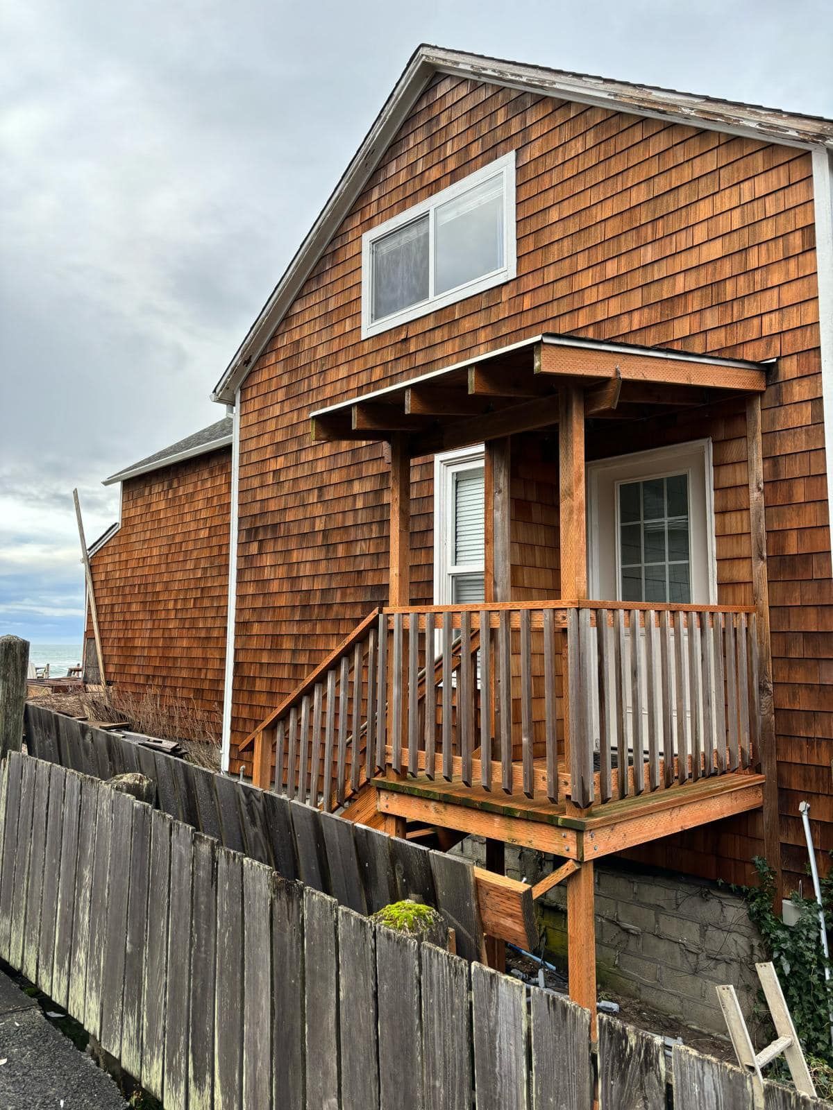 Wooden-shingled house with a porch and stairs. A weathered wooden fence runs along the side of the house. Overcast sky.