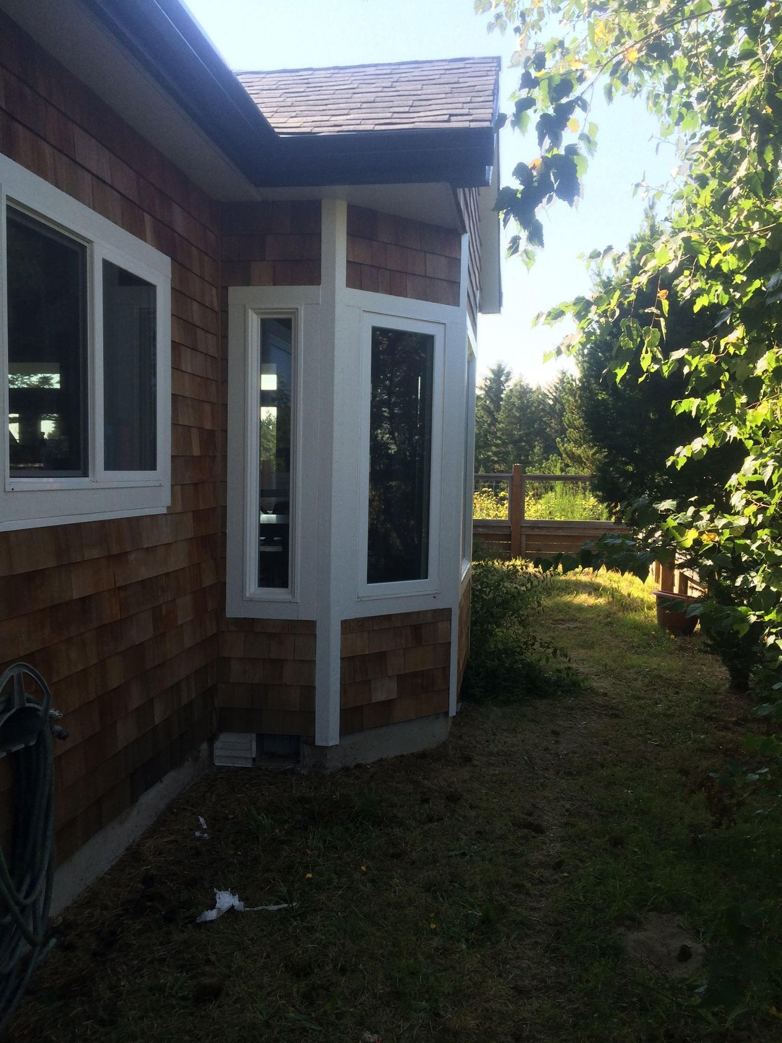 Exterior of a house with brown shingles, white trim, and a bay window. Green grass and trees surround it.