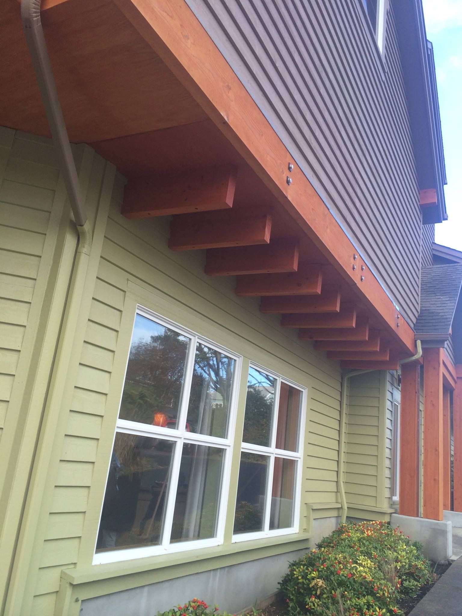 Green building with a brown wooden overhang and a large white-framed window; a bush is in front.