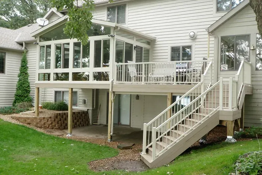 Beige two-story house with a screened porch and deck. Stairs lead to the deck. Green lawn and a small retaining wall in the foreground.