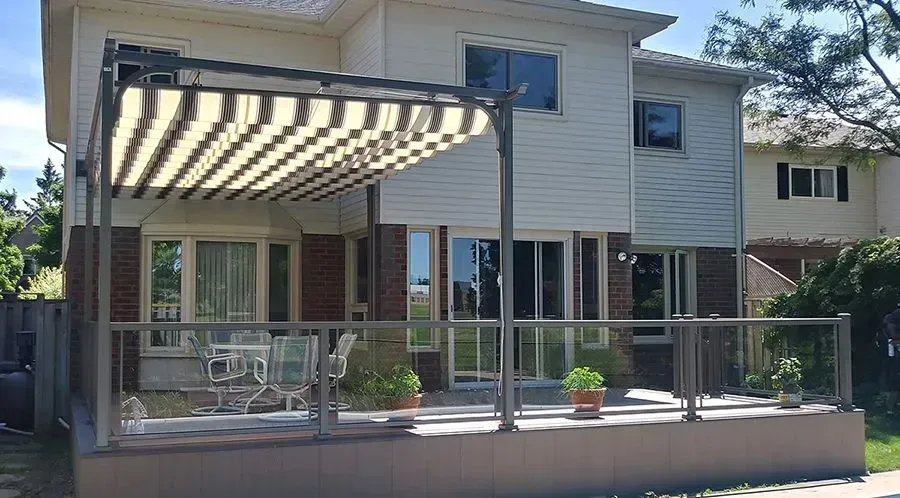 Back of a two-story house with a patio, pergola, and glass railings; striped awning.