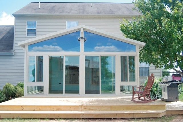 Sunroom addition on a wooden deck, with large glass windows reflecting the sky, next to a house and a grill.
