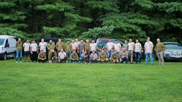 Group of men posing for a photo on a grassy lawn with trees in the background, vehicles parked nearby.