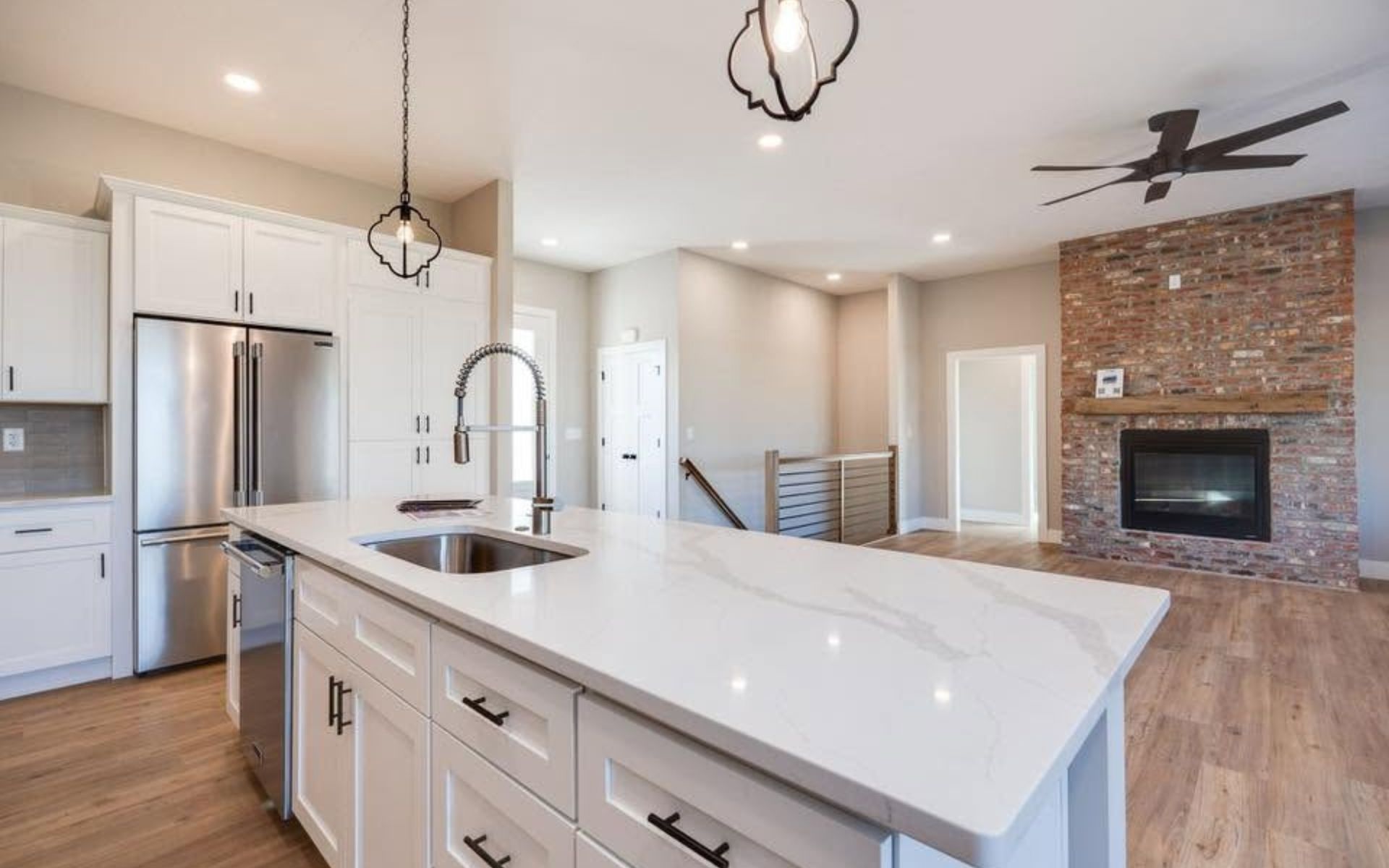 Modern open-concept kitchen and living room with white island, stainless fridge, brick fireplace, and wood floors
