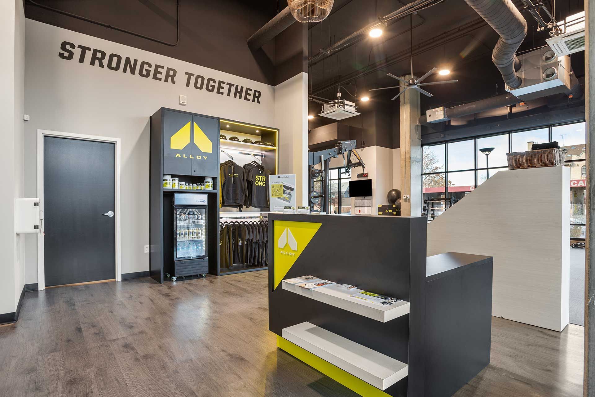 gym interior with a STRONGER TOGETHER sign, black and yellow reception desk, refrigerated drinks and apparel on display