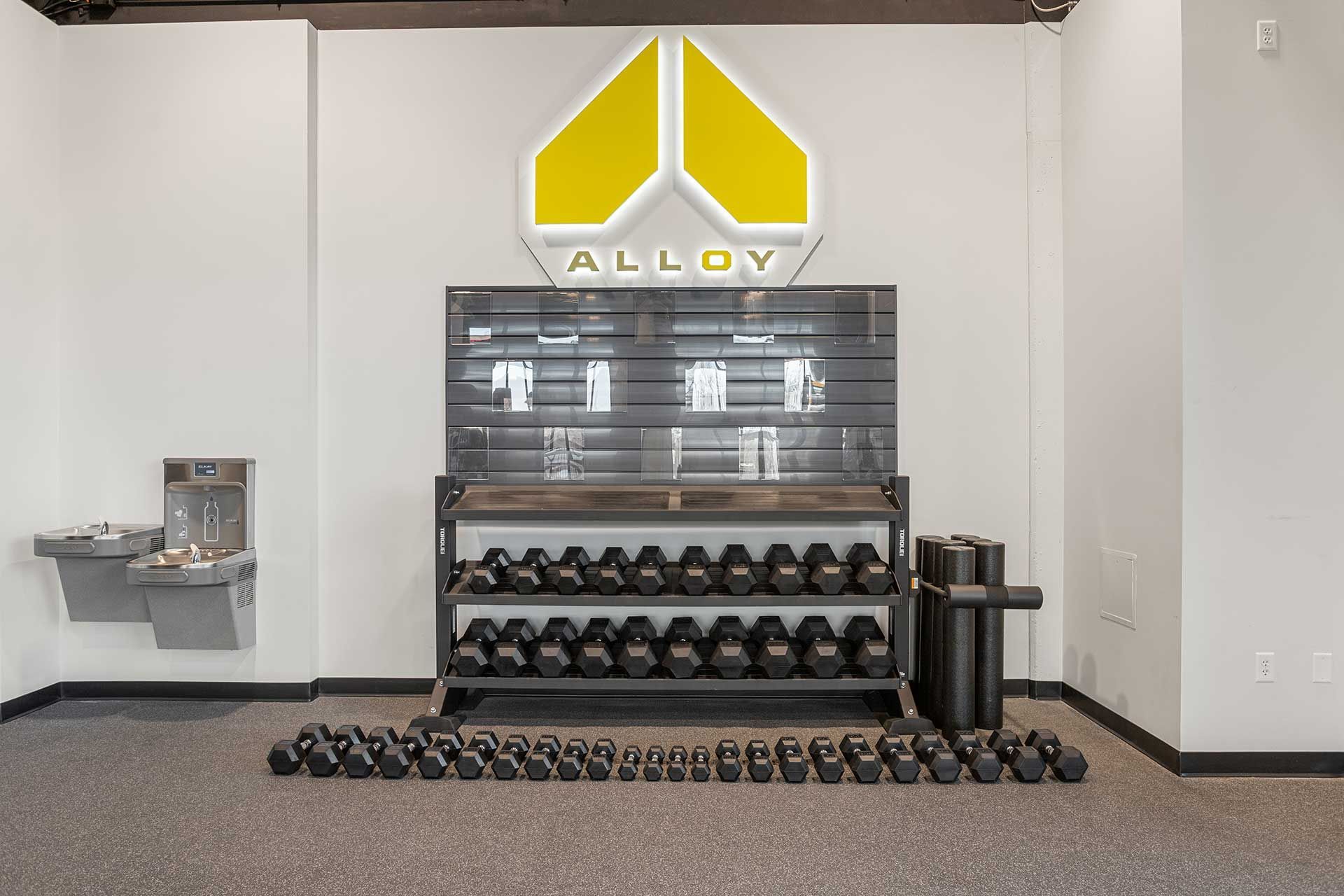 gym with dumbbell rack, water fountain, and yellow Alloy logo, dark weights on gray floor