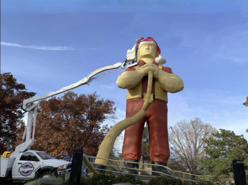 Large Paul Bunyan statue being worked on by a truck-mounted lift, wearing a Santa hat.