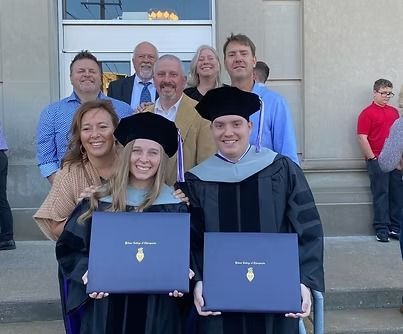 Two people in graduation gowns holding diplomas, surrounded by family, outside a building.