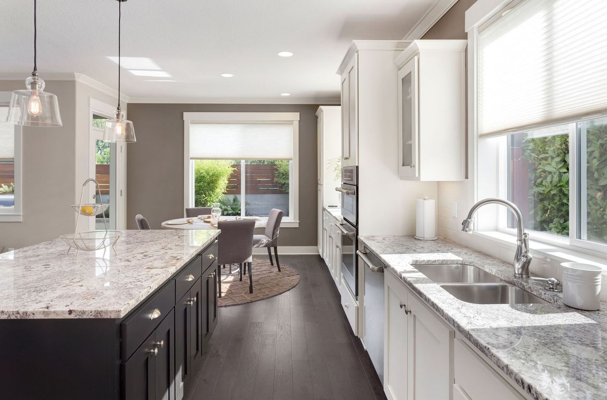 A modern kitchen featuring dark cabinets, a speckled granite island, white upper cabinetry, and a dining area in the back.