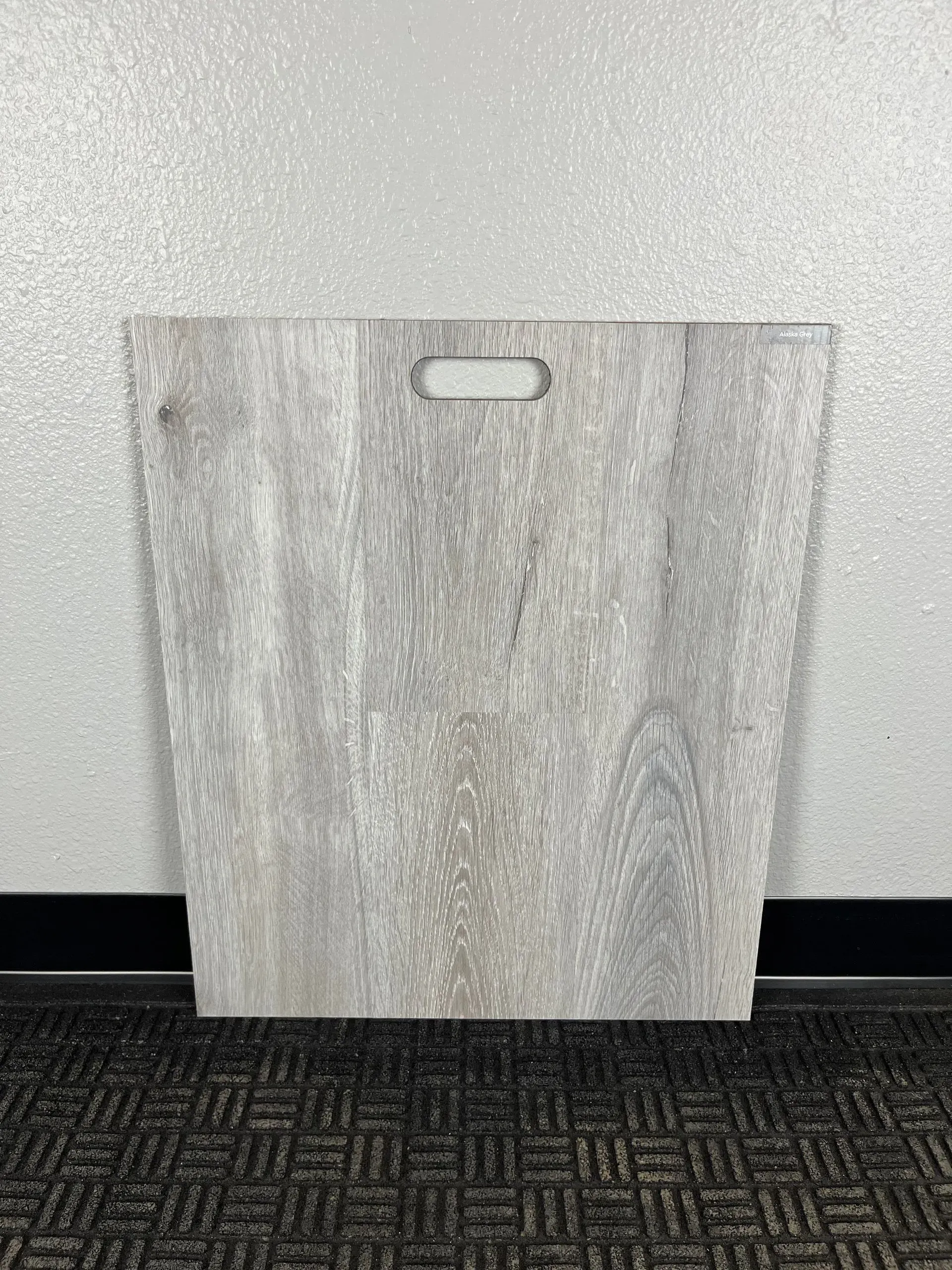 A square sample of light grey, wood-grain patterned flooring with a centered handle cutout, resting on a textured carpet.