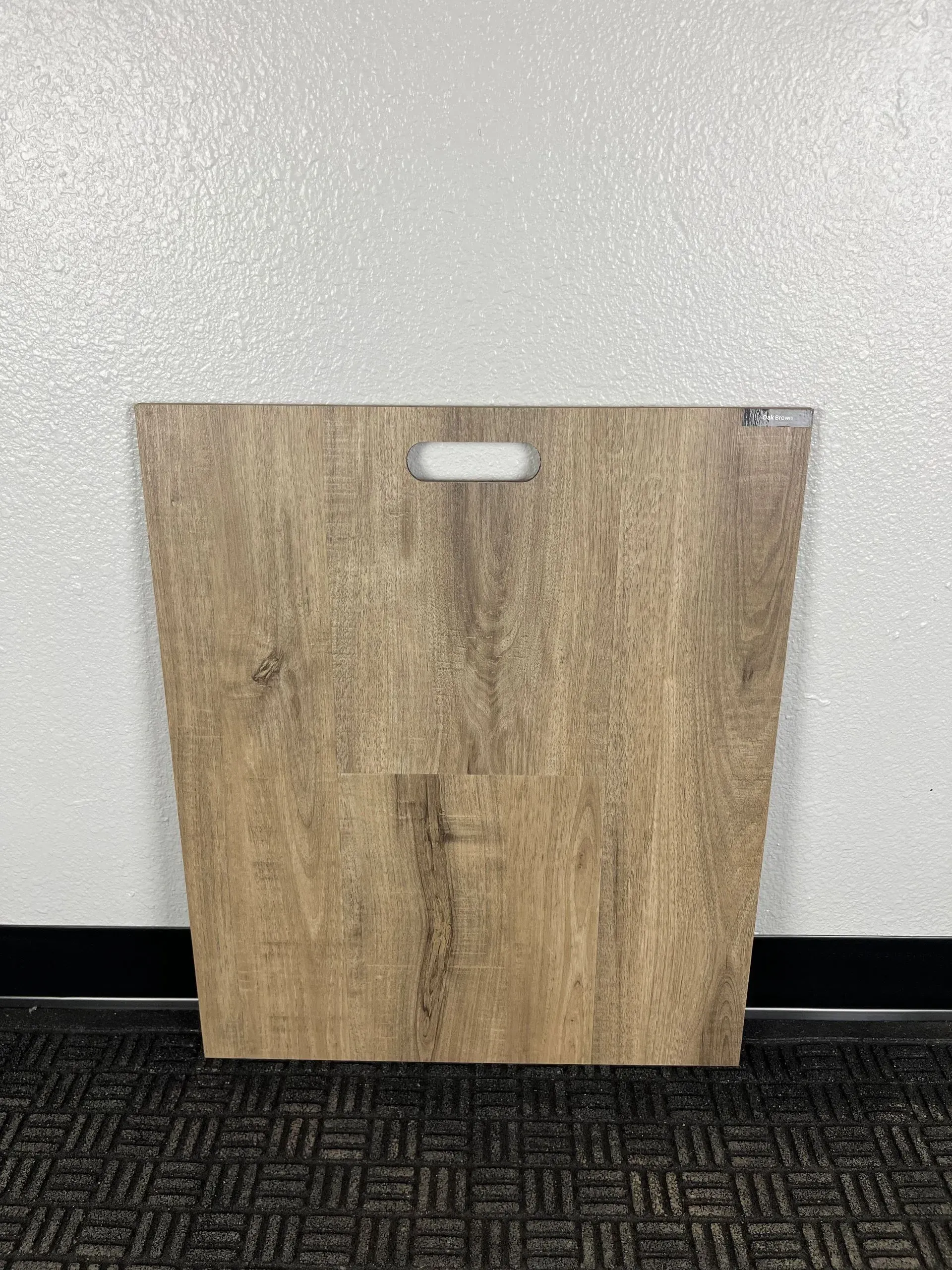 A rectangular wood-look flooring sample board with a cutout handle, standing against a white wall.