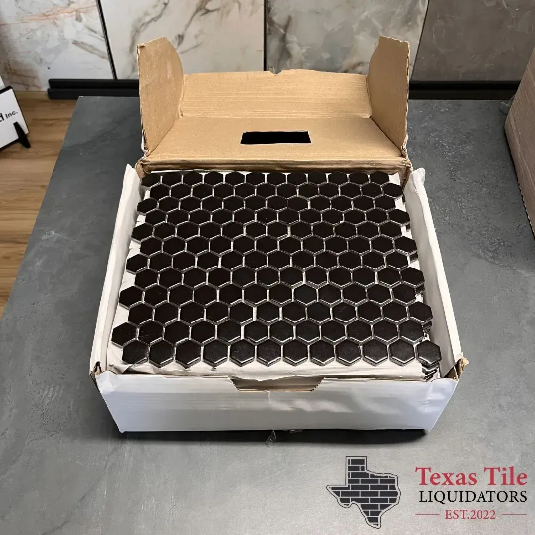 A box filled with black hexagonal mosaic tiles sitting on a gray surface, with a Texas Tile Liquidators logo below.