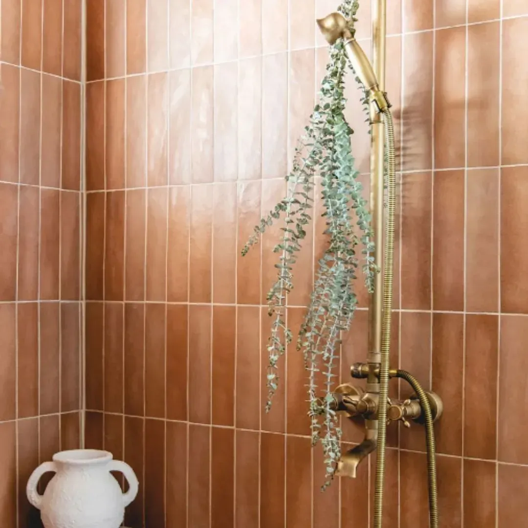 A brass shower fixture against warm-toned vertical tiles, with a hanging eucalyptus bunch and a white ceramic vase.