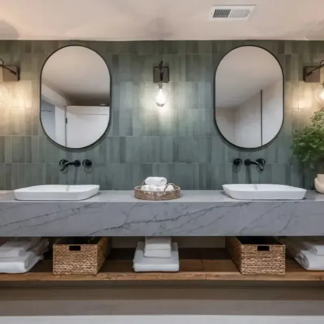 Double bathroom vanity with oval mirrors, dark green tiled wall, marble countertop, and open wood shelving with baskets.