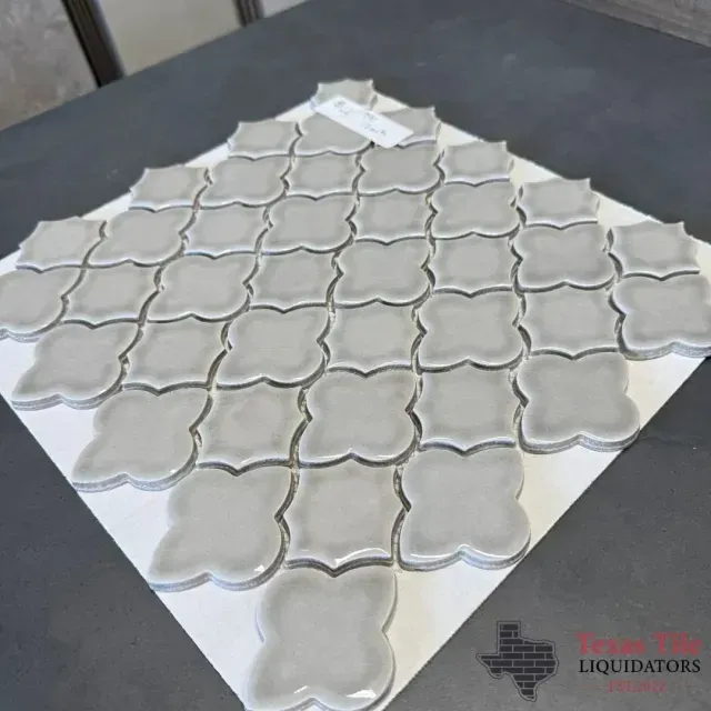 A sheet of light gray, four-lobed decorative tiles with a glossy finish arranged in a diamond pattern on a white backing.