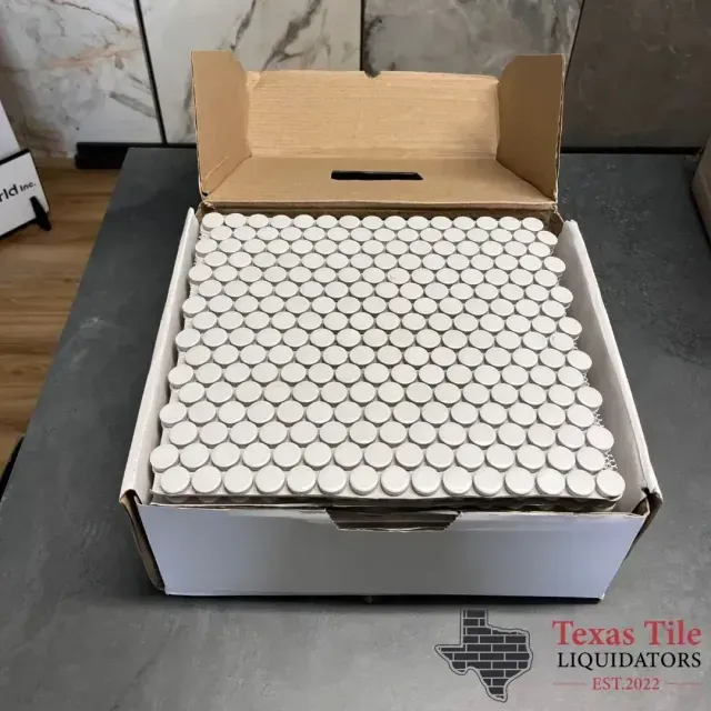 A box containing many white, circular penny tiles, positioned on a dark table, branded by Texas Tile Liquidators.