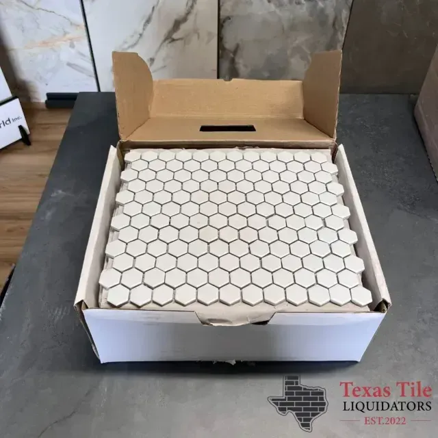 An open box containing white hexagonal mosaic tile sheets on a dark surface, with Texas Tile Liquidators branding.