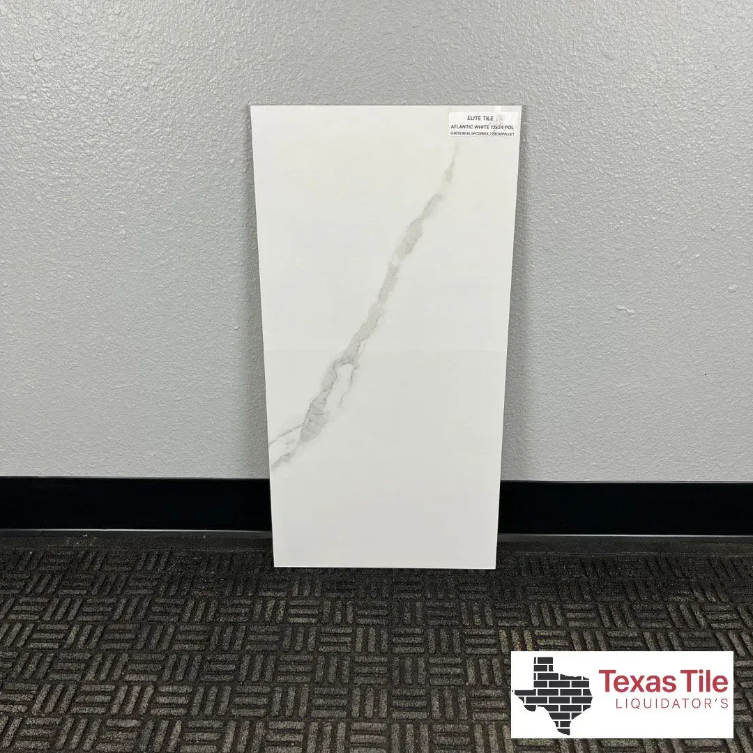 A white, rectangular floor tile with a single grey marble vein stands against a grey wall above a dark patterned carpet.