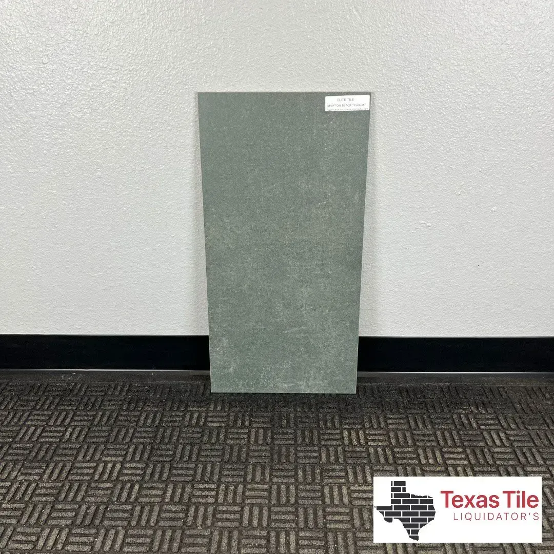 A rectangular, sage green stone tile sample stands upright against a white wall above a dark patterned carpet.