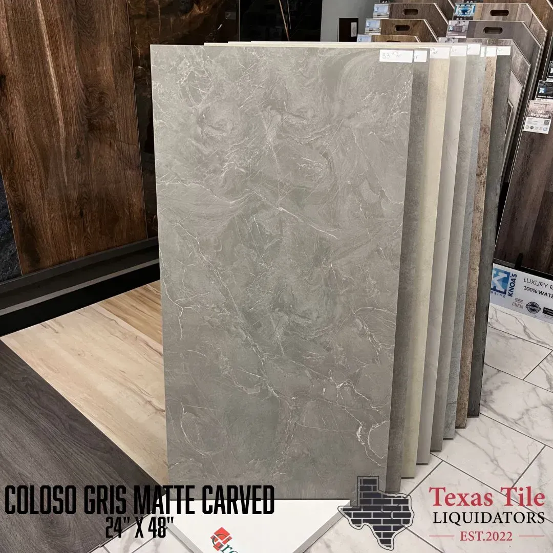 A showroom display of a large, rectangular, grey matte tile with a textured marble pattern, labeled Coloso Gris.