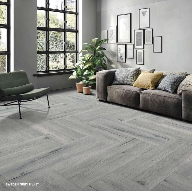 A modern living room with grey wood-look flooring, a plush sofa, an armchair, a potted plant, and wall-mounted art prints.