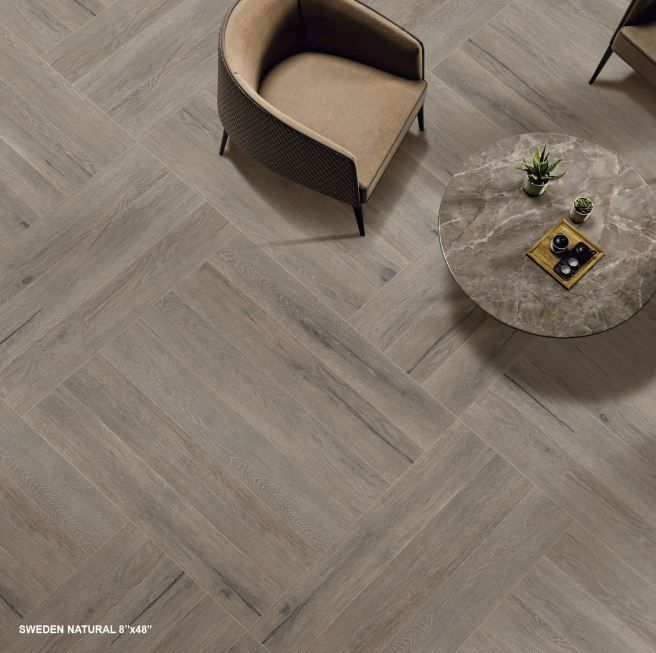 A top-down view of light wood-look floor tiles in a herringbone pattern, featuring an armchair and a round marble table.