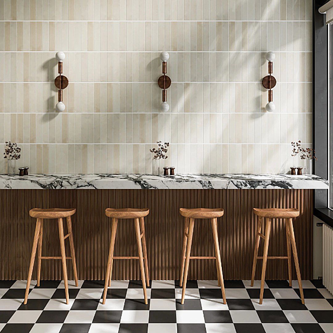 Four wooden stools line a marble-topped bar with wood-paneled sides against a light tile wall and black-and-white floors.