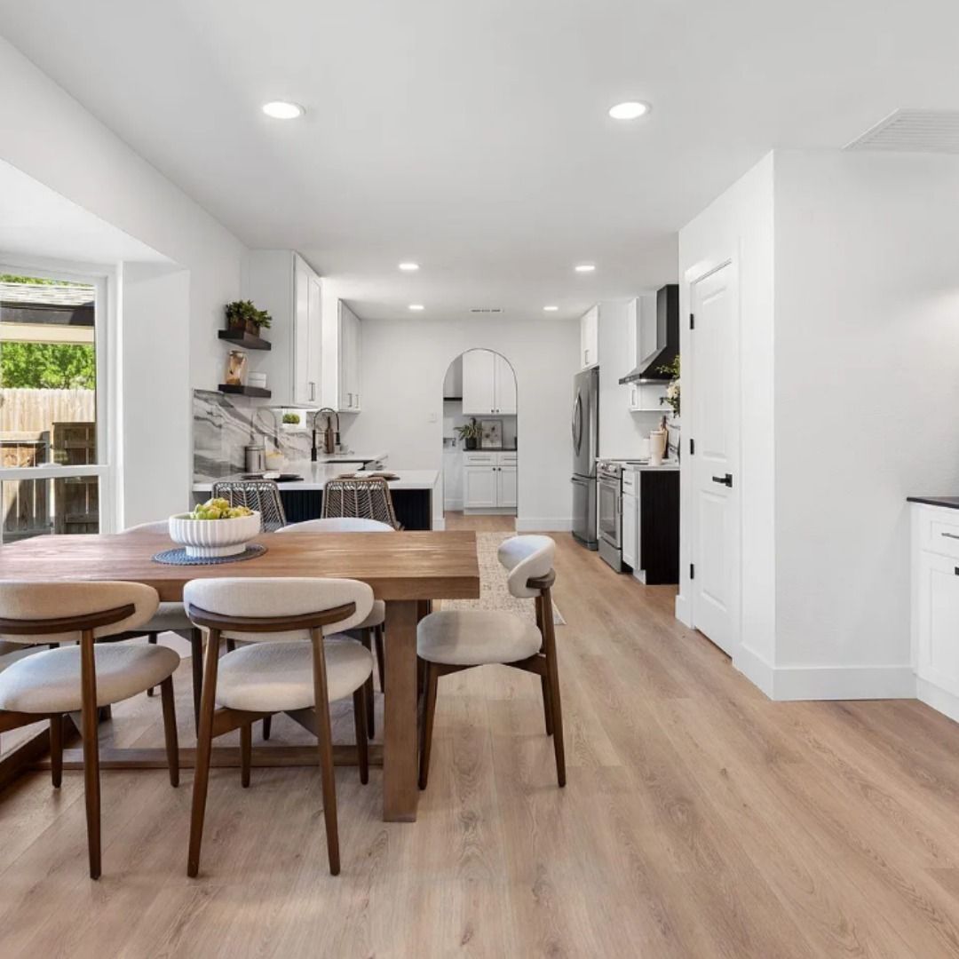 A bright, modern open-concept kitchen and dining area with white cabinets, wood flooring, and a wooden dining table.